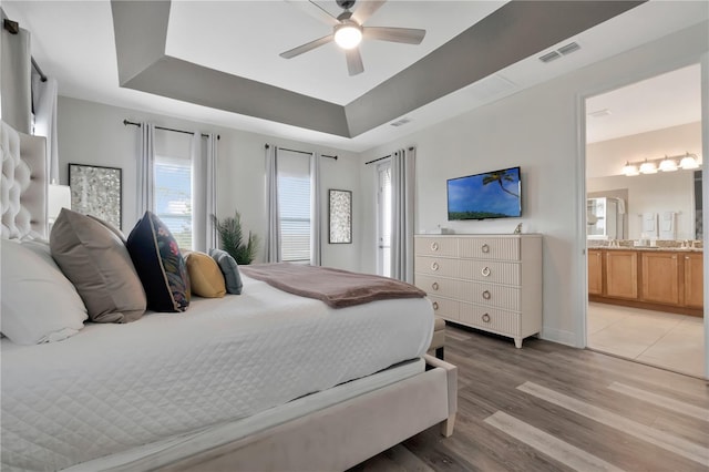 bedroom with ceiling fan, a raised ceiling, light wood-style flooring, ensuite bathroom, and visible vents