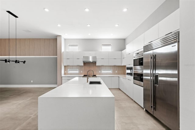 kitchen with stainless steel built in fridge, an island with sink, modern cabinets, wall chimney range hood, and tasteful backsplash