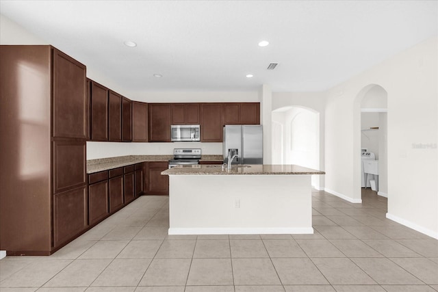 16780 Meadows St, Clermont, FL 34714 - kitchen with stainless steel appliances, arched walkways, light tile patterned floors, a center island with sink, and light stone counters