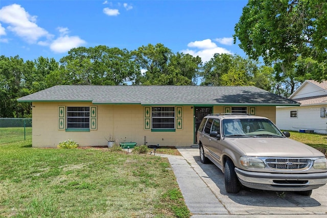 ranch-style home featuring a front lawn, a shingled roof, fence, and concrete block siding