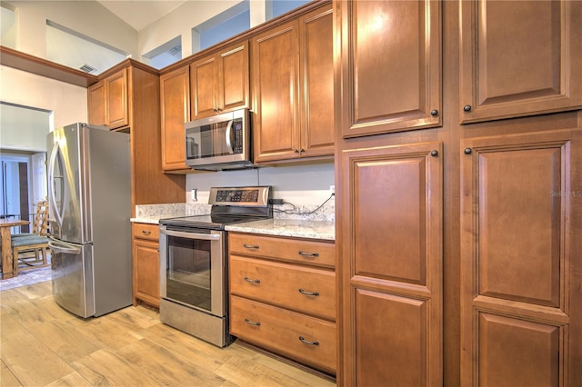kitchen featuring appliances with stainless steel finishes, light wood-style floors, brown cabinetry, and light stone countertops