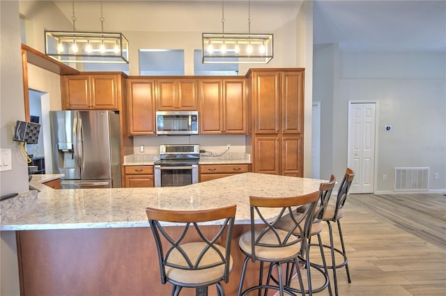 kitchen featuring appliances with stainless steel finishes, brown cabinets, light wood-type flooring, a towering ceiling, and a peninsula