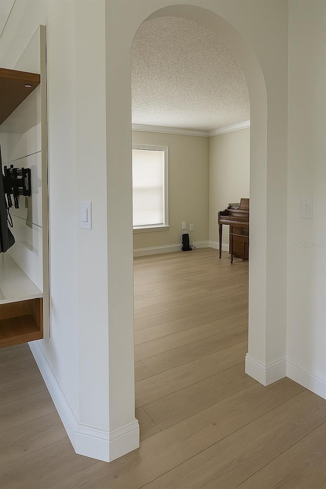 hall featuring crown molding, light wood-type flooring, a textured ceiling, baseboards, and arched walkways