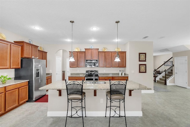 kitchen with brown cabinets, a breakfast bar, a center island with sink, arched walkways, and appliances with stainless steel finishes