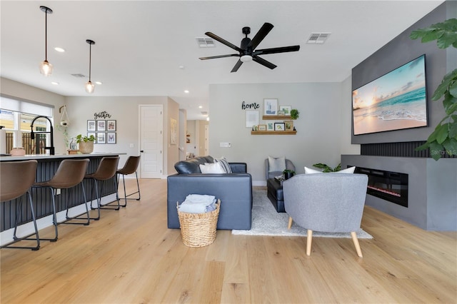 living area with ceiling fan, a glass covered fireplace, light wood-type flooring, and visible vents