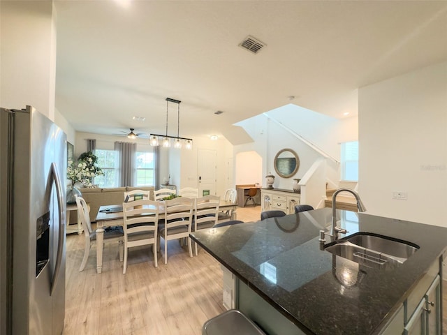 kitchen with stainless steel fridge with ice dispenser, a sink, a ceiling fan, light wood finished floors, and dark stone countertops