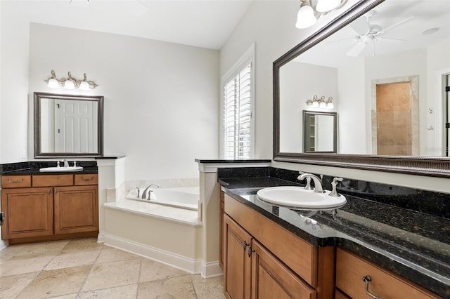 bathroom with a bath, a ceiling fan, two vanities, baseboards, and stone tile flooring