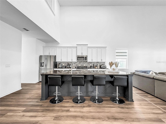 kitchen with wood finished floors, a towering ceiling, a kitchen island with sink, a kitchen bar, and appliances with stainless steel finishes