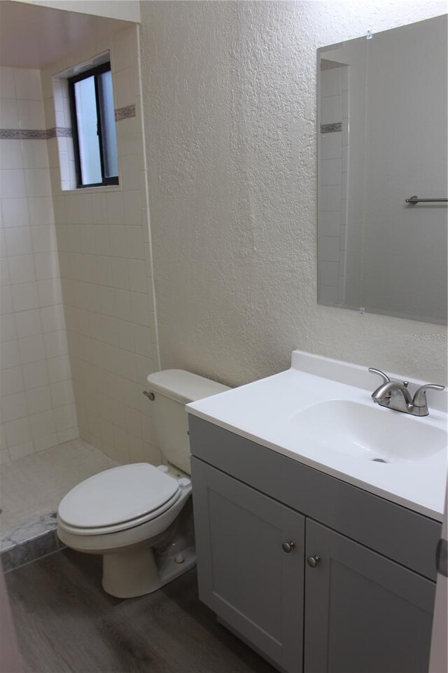 full bath with a textured wall, wood finished floors, toilet, vanity, and tiled shower
