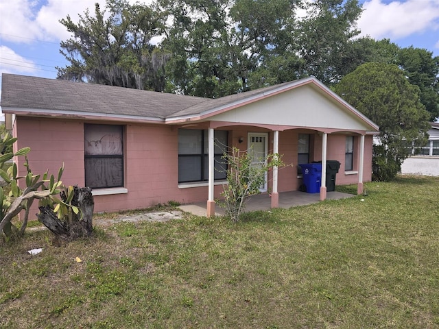 ranch-style home with concrete block siding, a front lawn, roof with shingles, and a patio area