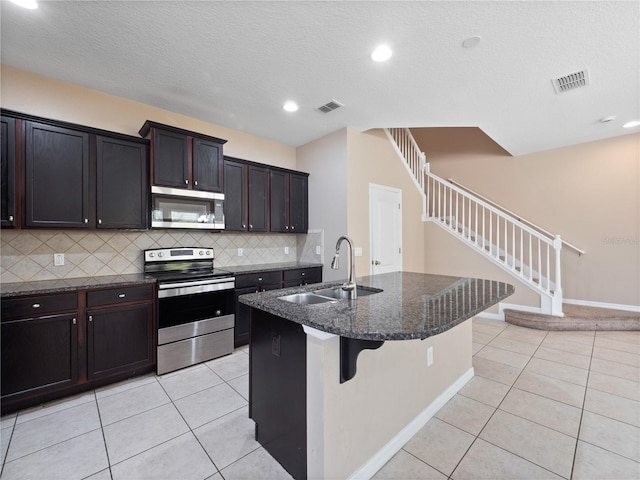 kitchen with stainless steel appliances, a sink, tasteful backsplash, light tile patterned flooring, and an island with sink