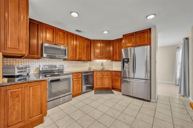 kitchen featuring appliances with stainless steel finishes, light tile patterned floors, tasteful backsplash, dark stone counters, and a sink
