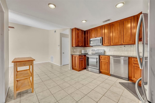 kitchen with stainless steel appliances, backsplash, light tile patterned floors, dark stone counters, and brown cabinetry