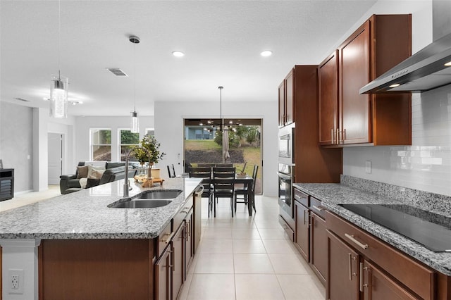 kitchen with wall chimney range hood, black electric cooktop, a sink, oven, and an island with sink
