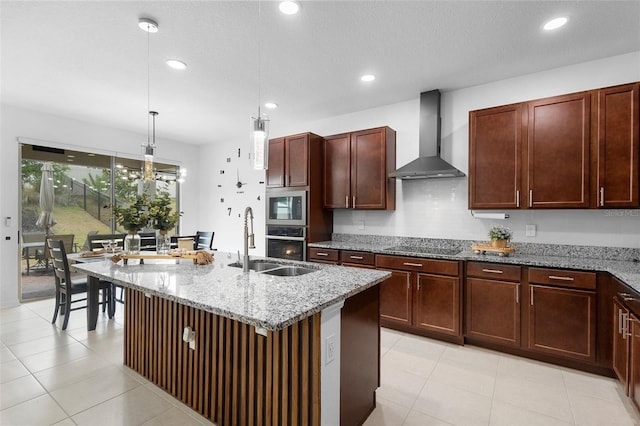 kitchen featuring wall chimney exhaust hood, appliances with stainless steel finishes, light stone counters, recessed lighting, and a kitchen island with sink