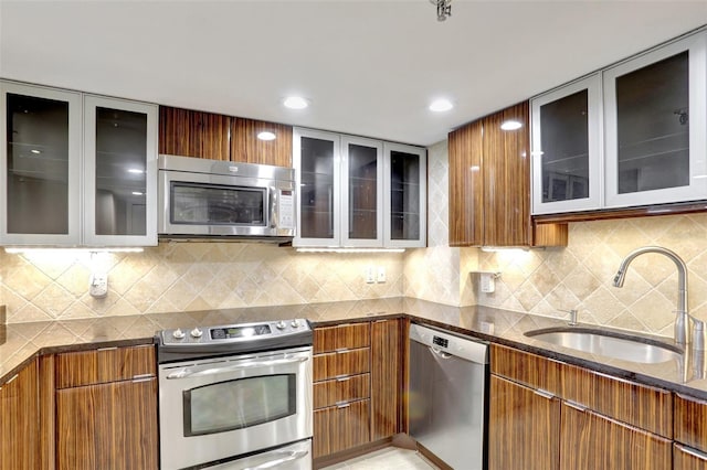 kitchen featuring stainless steel appliances, a sink, dark stone countertops, glass insert cabinets, and brown cabinets