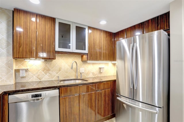 kitchen featuring appliances with stainless steel finishes, a sink, dark stone countertops, tasteful backsplash, and glass insert cabinets