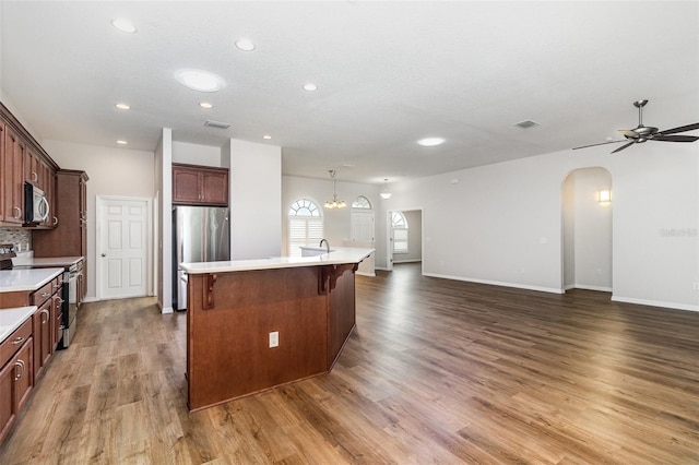 kitchen with appliances with stainless steel finishes, arched walkways, light countertops, wood finished floors, and a kitchen island with sink