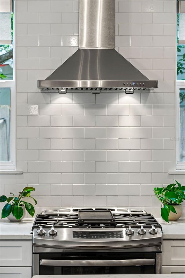 kitchen featuring wall chimney exhaust hood, stainless steel gas range oven, decorative backsplash, and white cabinets