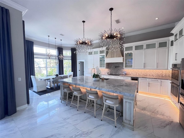 kitchen with a chandelier, tasteful backsplash, ornamental molding, white cabinets, and a large island with sink