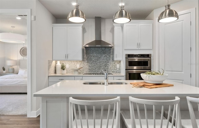 kitchen featuring wall chimney exhaust hood, a sink, wood finished floors, double oven, and backsplash