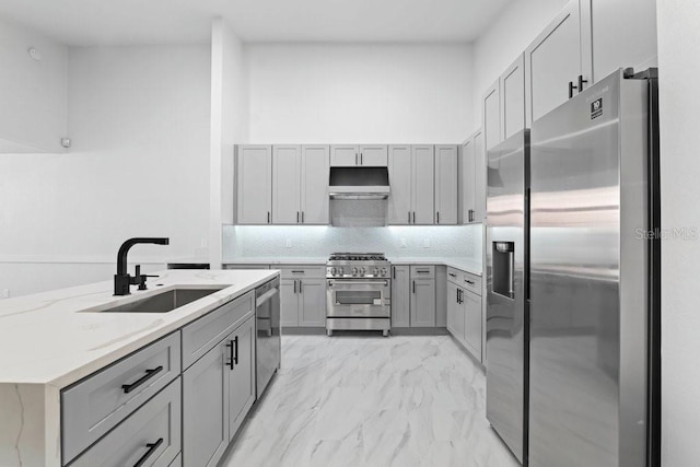 kitchen with stainless steel appliances, gray cabinetry, a sink, light marble finish floors, and under cabinet range hood