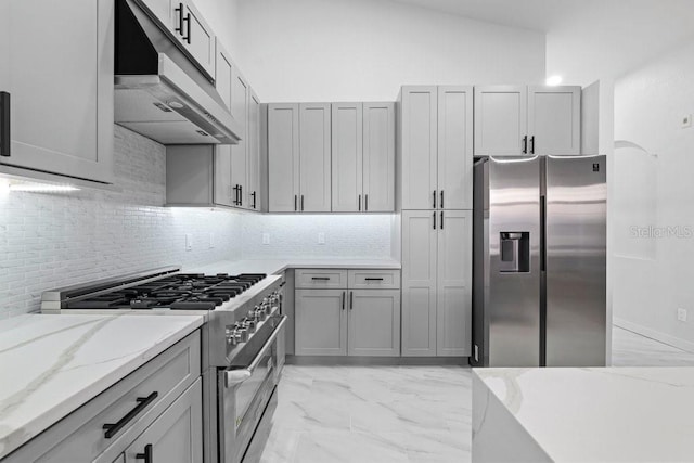 kitchen featuring appliances with stainless steel finishes, under cabinet range hood, tasteful backsplash, light marble finish flooring, and light stone counters