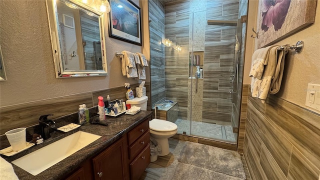 full bath with a textured wall, a stall shower, toilet, vanity, and a wainscoted wall