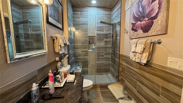 bathroom featuring toilet, a stall shower, a textured wall, recessed lighting, and a wainscoted wall