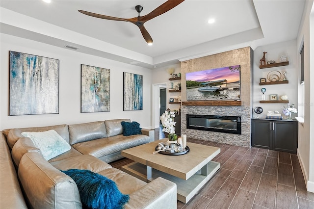 living area featuring wood finish floors, a tray ceiling, a ceiling fan, a large fireplace, and recessed lighting