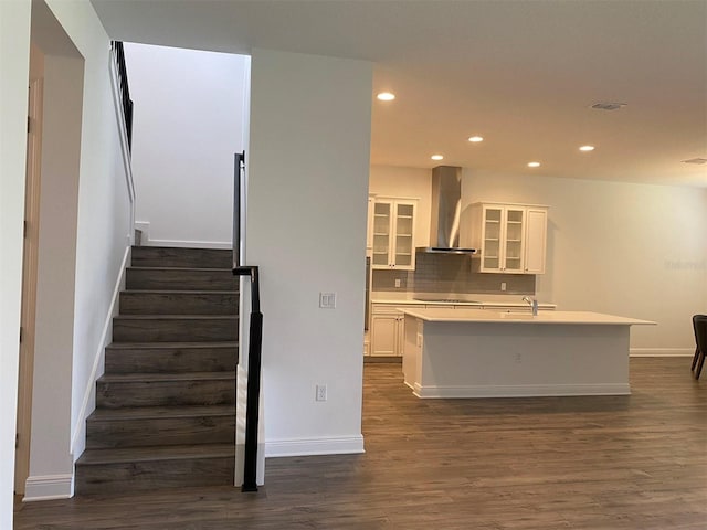 kitchen featuring wall chimney range hood, dark wood-type flooring, recessed lighting, baseboards, and a kitchen island with sink