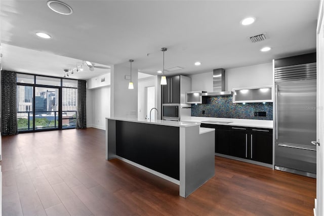 kitchen with wall chimney range hood, built in fridge, tasteful backsplash, light countertops, and dark cabinetry