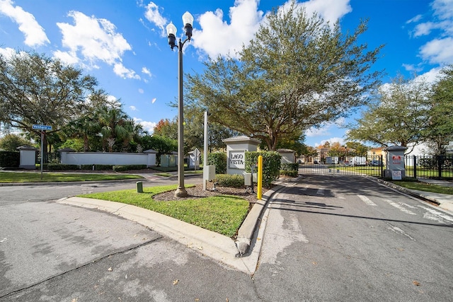 view of asphalt road featuring curbs, a gate, street lighting, and sidewalks