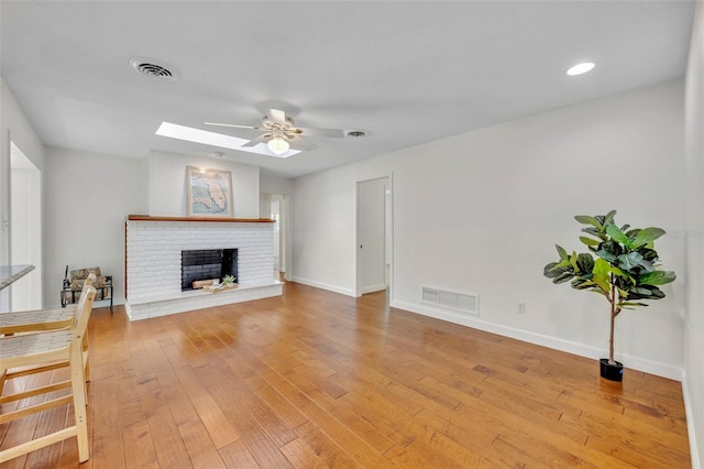 living room featuring ceiling fan, a skylight, a brick fireplace, light wood-style floors, and baseboards