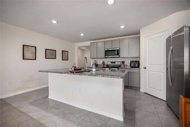 kitchen featuring stainless steel appliances, gray cabinetry, a center island with sink, light tile patterned flooring, and dark stone counters