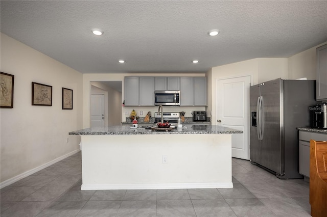 kitchen with appliances with stainless steel finishes, gray cabinetry, dark stone countertops, an island with sink, and light tile patterned floors