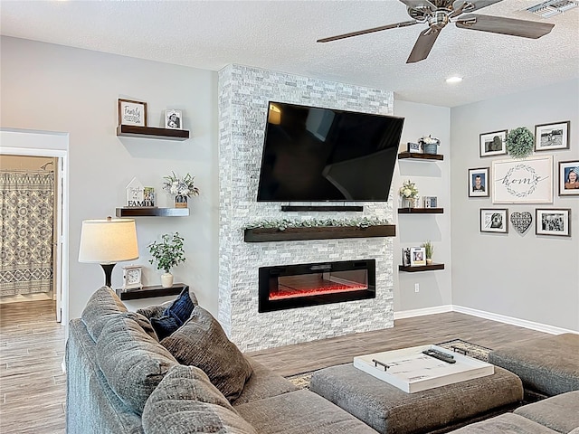 living area featuring wood finished floors, ceiling fan, a stone fireplace, a textured ceiling, and baseboards