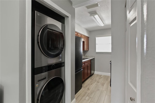 clothes washing area with stacked washer / drying machine, light wood-style flooring, baseboards, and a textured ceiling
