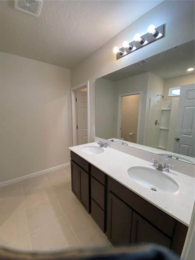 bathroom featuring a shower, double vanity, tile patterned floors, baseboards, and a textured ceiling