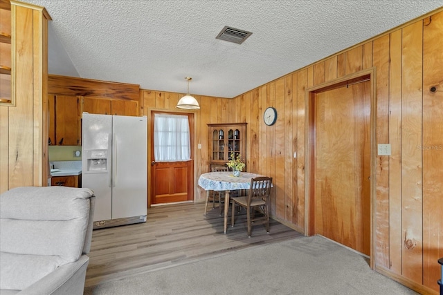 kitchen with white refrigerator with ice dispenser, wooden walls, brown cabinetry, a textured ceiling, and light colored carpet