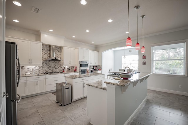 kitchen with stainless steel appliances, wall chimney range hood, an island with sink, ornamental molding, and decorative backsplash