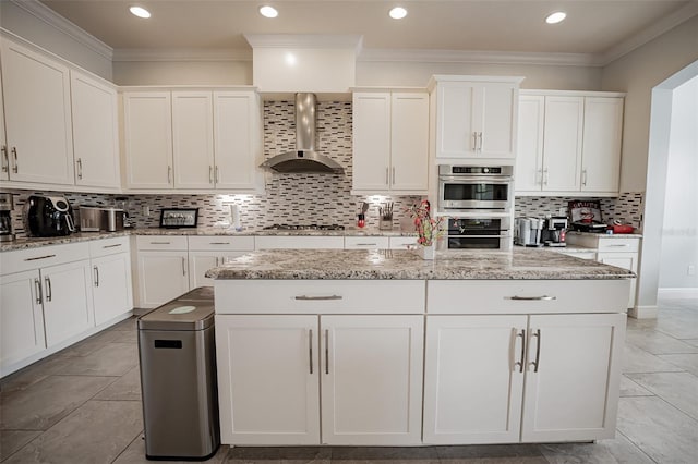kitchen with wall chimney range hood, appliances with stainless steel finishes, backsplash, white cabinets, and a kitchen island