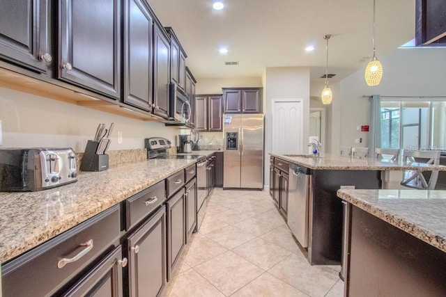 kitchen featuring appliances with stainless steel finishes, light stone counters, hanging light fixtures, light tile patterned flooring, and an island with sink