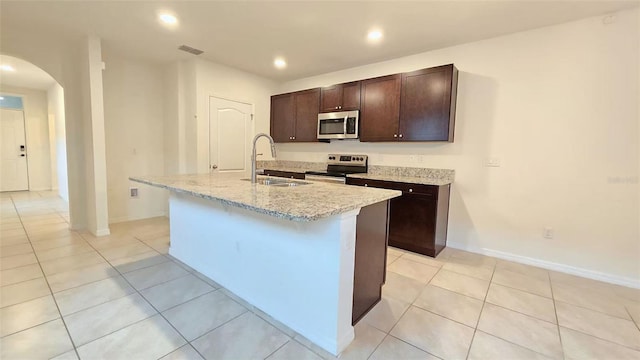 3202 Timber Hawk Cir, Ocoee, FL 34761 - kitchen featuring stainless steel appliances, arched walkways, a center island with sink, dark brown cabinets, and light tile patterned flooring