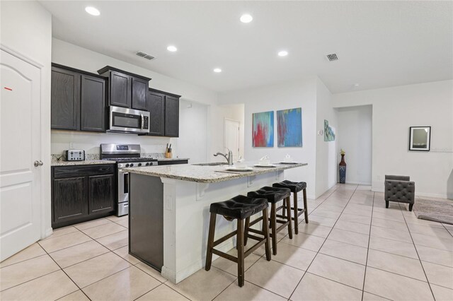 7033 Oakwood St, Davenport, FL 33837 - kitchen featuring stainless steel appliances, light tile patterned floors, light stone counters, a kitchen island with sink, and a breakfast bar area