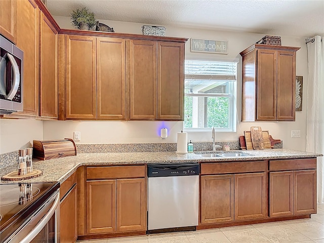 kitchen with appliances with stainless steel finishes, a sink, light stone countertops, brown cabinetry, and light tile patterned flooring