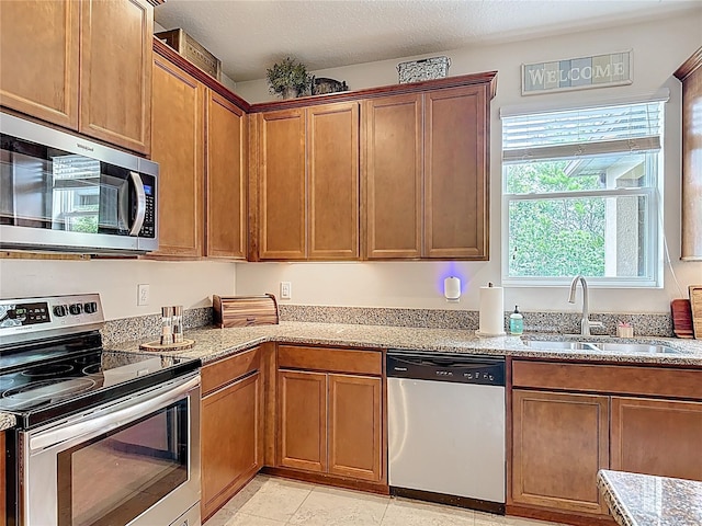 kitchen featuring stainless steel appliances, a sink, light stone counters, and brown cabinetry