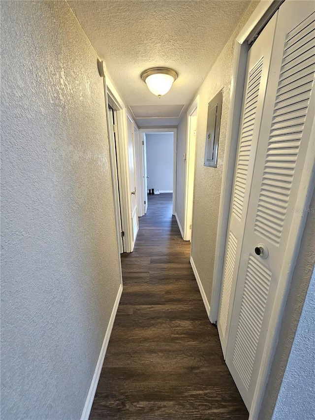 corridor featuring a textured wall, wood finished floors, baseboards, a textured ceiling, and electric panel