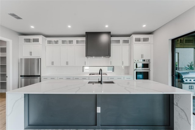 15942 Vetta Dr, Bella Collina, FL 34756 - kitchen featuring appliances with stainless steel finishes, ventilation hood, a large island with sink, light stone countertops, and white cabinetry