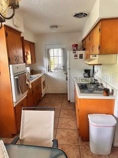kitchen with ventilation hood, light tile patterned flooring, brown cabinetry, appliances with stainless steel finishes, and light countertops